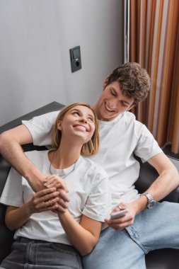 happy young travelers holding hands and looking at each other on couch in hotel apartments