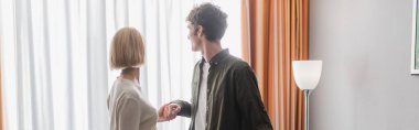 brunette man and blonde woman holding hands near window in hotel room, banner