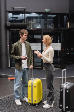 young man with mobile phone smiling at blonde girlfriend talking near travel bags in hotel lobby