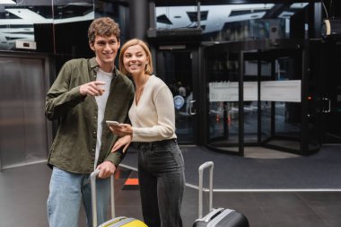 young man pointing with finger and looking away near smiling girlfriend and travel bags in hotel