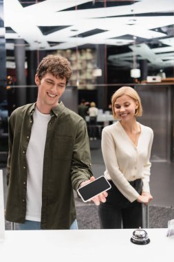 happy man holding smartphone with blank screen near reception and girlfriend in hotel