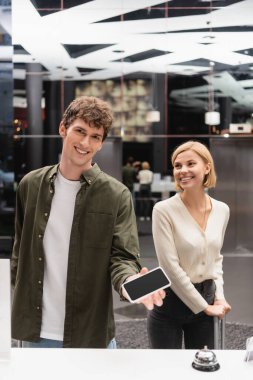 young man holding smartphone with blank screen near reception counter and happy blonde girlfriend in hotel