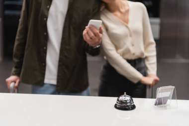 service bell and card holder on reception desk near cropped couple with smartphone on blurred background