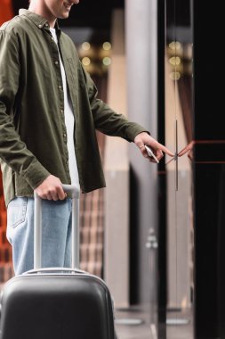 partial view of smiling traveler with baggage and smartphone standing near doors of elevator and pressing call button
