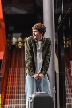 cheerful young man with suitcase standing in hotel lobby and looking away