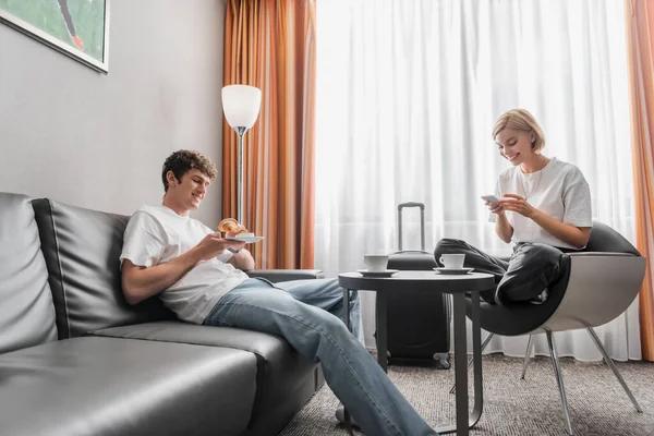 stock image smiling woman using smartphone in armchair near boyfriend with croissant on couch in hotel room