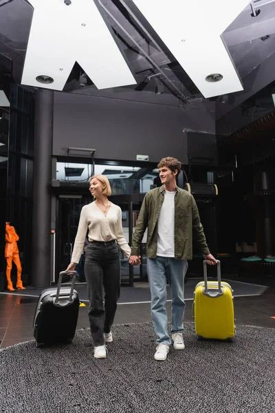 full length of cheerful couple holding hands and looking away while walking with suitcases in lobby of modern hotel