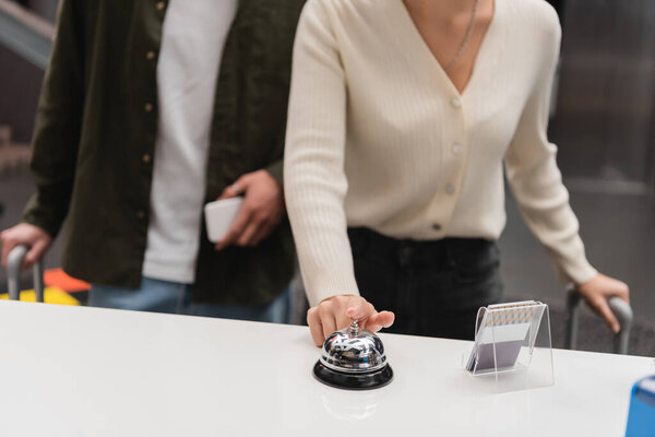 partial view of woman ringing service bell on reception near blurred boyfriend with smartphone in hotel