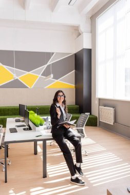 full length of manager in black suit holding notebook near desk with computers in spacious office