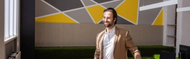 young corporate manager in beige blazer smiling and looking away in office, banner