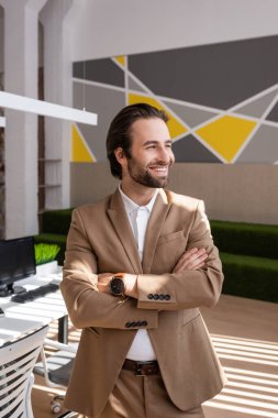 young and happy businessman in beige blazer standing with crossed arms and looking away in office