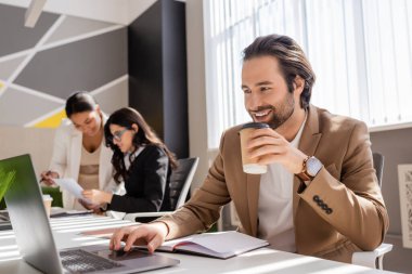 smiling manager with takeaway drink looking at laptop near multiethnic businesswomen on blurred background