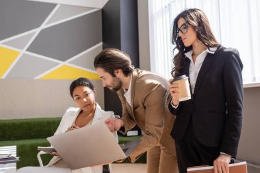 young manager showing laptop to multiracial businesswoman near colleague standing with coffee to go