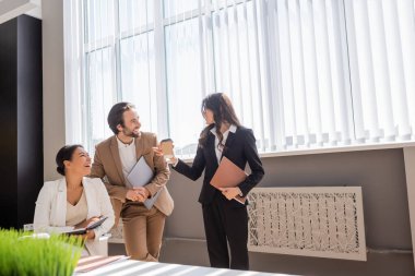 businesswoman with folder and takeaway drink talking to cheerful colleagues near large window in office