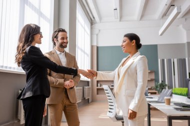 young and smiling manager looking at interracial businesswomen shaking hands in office