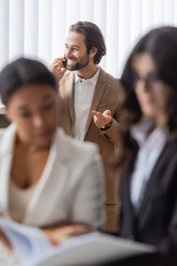 smiling businessman gesturing and talking on mobile phone near interracial colleagues on blurred foreground