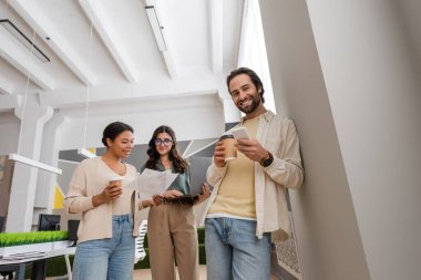 manager with paper cup and smartphone looking at camera near interracial businesswomen on background