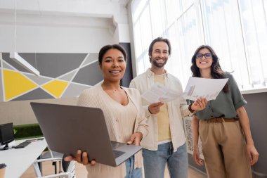 happy multiethnic business colleagues with laptop and documents smiling at camera in office