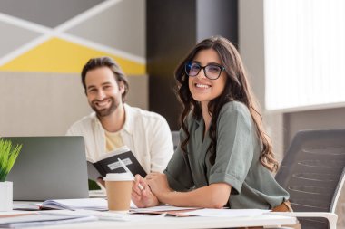 happy manager in eyeglasses smiling at camera near blurred colleague and paper cup on work desk