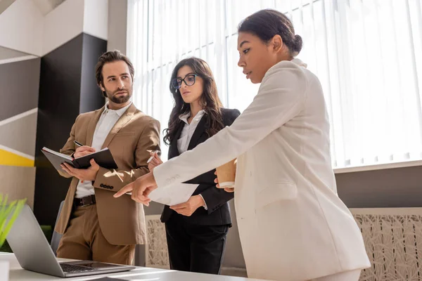 multiracial businesswoman pointing at laptop near serious managers with notebook and document