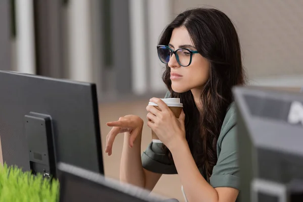 thoughtful businesswoman in eyeglasses holding coffee to go and pointing at computer monitor while working in office