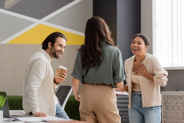 stock image excited multiracial businesswoman touching chest while laughing near colleagues in office