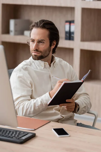 young businessman in eyeglasses looking at blurred monitor and writing in notebook while working in office