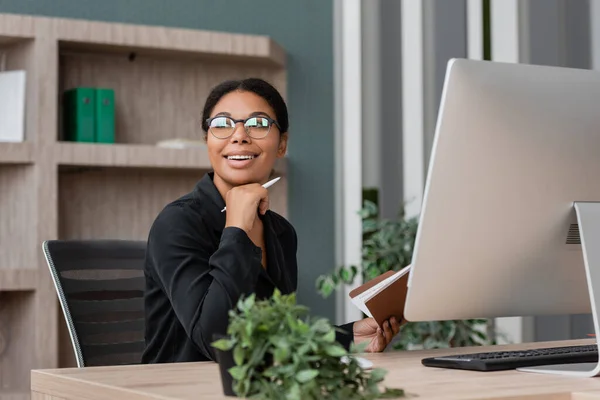 stock image cheerful multiracial businesswoman in eyeglasses looking away near computer monitor and blurred potted plant
