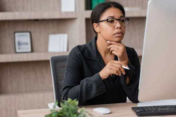 stock image thoughtful multiracial manager in eyeglasses holding pen and looking at computer monitor in office