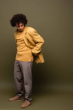 Sarı ceketli ve pantolonlu Afro-Amerikan bir adam. Gri yeşil arka planda eli arkadan poz veriyor.