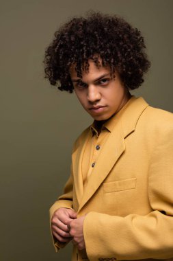 Kıvırcık esmer saçlı, sarı ceketli ve gri renkli kameraya bakan Afro-Amerikan genç adam.