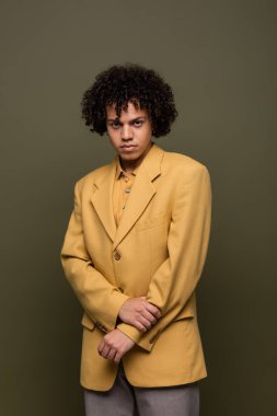 Esmer Afrikalı Amerikalı trend sarı ceketli adam zeytin grisi arka planda kameraya bakıyor.