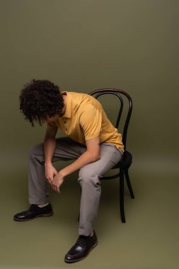 Sarı polo tişörtlü, siyah deri ayakkabılı, başı eğilmiş, gri arka planda oturan Afro-Amerikan bir adam.