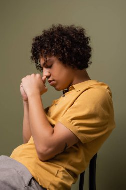 Sarı polo tişörtlü Afro-Amerikan bir adamın yan görüntüsü. Sandalyeye oturmuş, gözleri kapalı ve elleri yüzü gri yeşil arka planda.