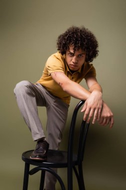 Dövmeli Afro-Amerikan adam, sarı polo gömlekli, gri yeşil arka plandaki kameraya bakarken sandalyeyle poz veriyor.