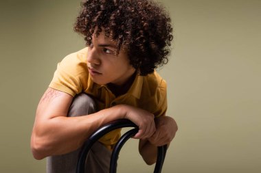 Kıvırcık saçlı şık Afro-Amerikalı adam, gri sandalyenin yanında poz verirken gözlerini kaçırıyor.