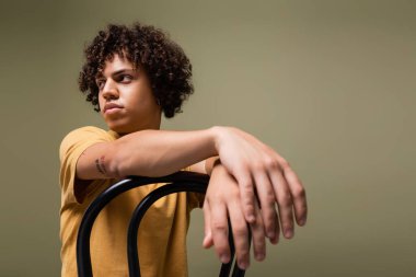 Kıvırcık esmer saçlı genç Afrikalı Amerikalı adam gri sandalyede tek başına otururken gözlerini kaçırıyor.