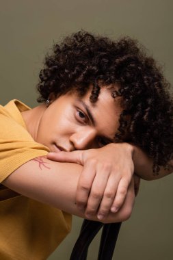 Esmer Afrikalı Amerikalı adam sandalyeye yaslanıyor ve gri renkli kameraya bakıyor.