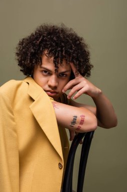 Dövmeli Afro-Amerikalı adam, sarı ceketli ve yüzünün yanında oturan gri tenli.