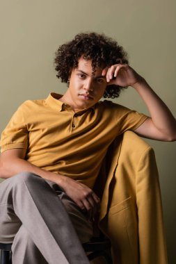 Sarı polo tişörtlü modaya uygun Afro-Amerikan adam bacak bacak üstüne atmış ve başı gri arka planda oturuyor.