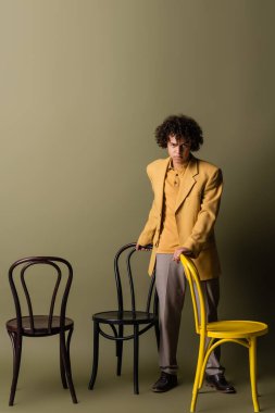 Tam boy, siyah ve sarı koltukların yanında, zeytin grisi arka planda şık giyinmiş, Afro-Amerikan bir adam.