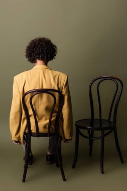 Esmer, Afro-Amerikalı, kıvırcık saçlı, sarı ceketli, gri arka planda siyah koltukta oturan adam.