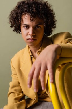 Sarı ceketli şık Afro-Amerikan adam gri önplanda bulanık ön planda oturuyor ve kameraya bakıyor.