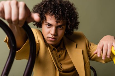 Gri renkli koltukların yakınındaki kameralara bakan modaya uygun giyinmiş Afro-Amerikan genç adam.