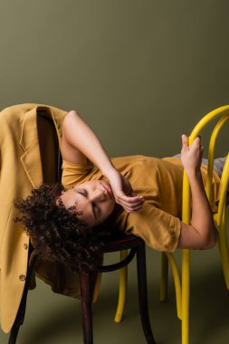 Genç Afro-Amerikalı, gözleri kapalı, gri yeşil arka planda sarı ceketin yanındaki sandalyelere uzanmış.