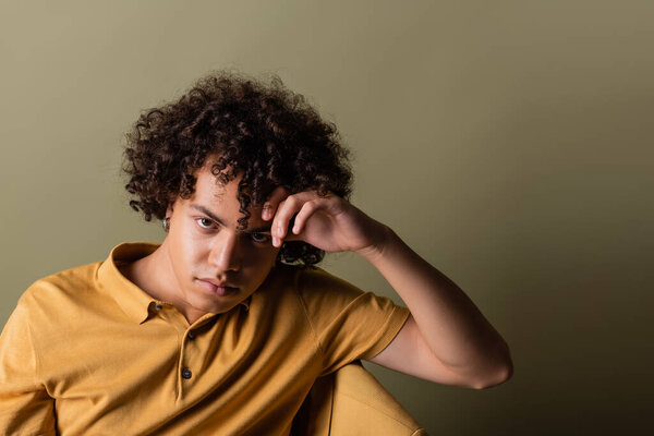 curly african american man in yellow polo shirt holding hand near head and looking at camera while sitting on grey green background