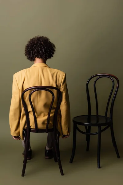 back view of brunette african american man with curly hair sitting in yellow blazer on black chair on grey green background