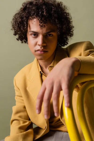 Sarı ceketli şık Afro-Amerikan adam gri önplanda bulanık ön planda oturuyor ve kameraya bakıyor.