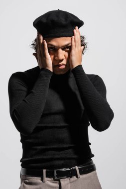 Siyahi balıkçı yaka kazaklı ve bereli şık Afro-Amerikan adam yüzünün yanında el ele tutuşmuş ve gri renkli kameraya bakıyor.