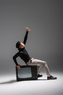 Afrika kökenli Amerikalı bir adam, modaya uygun giyinmiş, klasik TV setinde oturuyor ve gri arka planda elini kaldırıyor.